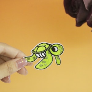 May include: A green and white embroidered iron-on patch of a cartoon sea turtle with a smiling face.