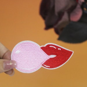 Könnte beinhalten: Ein gestickter Patch mit einem rosa Kaugummi, der von einer roten Lippe angeblasen wird.
