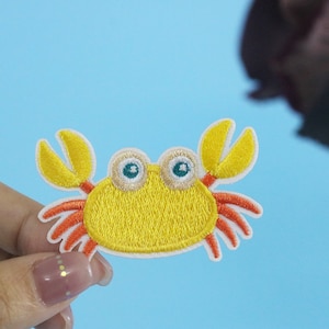 May include: A yellow embroidered crab with orange claws and white eyes. The crab is on a blue background.