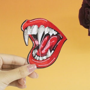 Pode incluir: Etiqueta bordada vermelha de uma boca grande aberta com dentes afiados e uma língua longa e vermelha. A boca está bem aberta, mostrando os dentes e a língua.