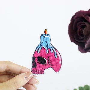 Puede incluir: Cráneo rosa con una vela azul en la parte superior. La vela se está derritiendo y goteando cera. El cráneo es un parche.