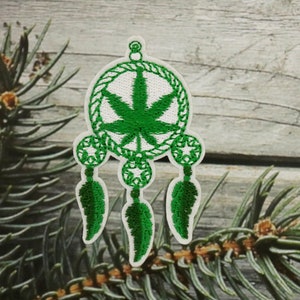 以下が含まれることがあります： 緑の葉っぱが刺繍された白いドリームキャッチャーに、緑の羽根が3本ついています。