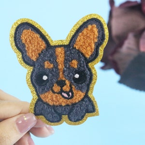 Op de afbeelding: Een geborduurde hondenpatch in zwart en bruin met een gouden glitterrand. De hond heeft een vrolijke uitdrukking met een roze tong die uitsteekt.