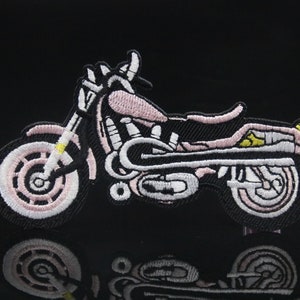 以下が含まれることがあります： 白と黄色のディテールが付いたピンクの刺繍のオートバイのパッチ。パッチは黒い背景です。