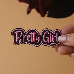 May include: A black embroidered patch with the words "Pretty Girl" in pink script. The patch has a pink border and is held against a peach-colored background.