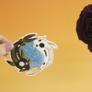 May include: A black and white yin yang fish design embroidered patch with blue and gold accents. The patch is held by a hand against a light orange background.