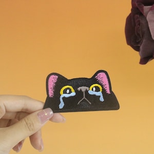 May include: Black embroidered iron-on patch of a cat with pink ears and yellow eyes, crying blue tears. The cat is looking directly at the viewer.