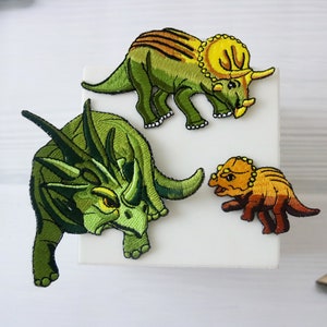 May include: Embroidered dinosaur patches in shades of green, yellow, and brown. The patches depict a Triceratops and other dinosaur species. The patches are on a white surface.