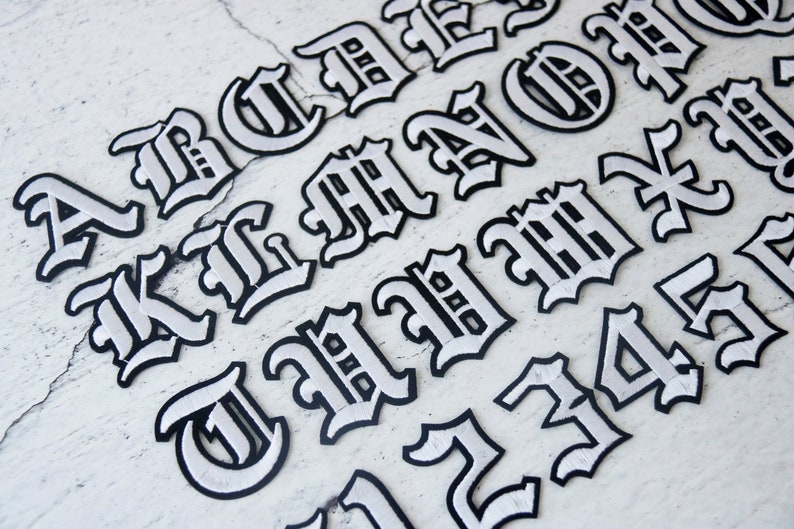 May include: White iron-on letters in a gothic font style. The letters are A, B, C, D, E, F, G, H, I, J, K, L, M, N, O, P, Q, R, S, T, U, V, W, X, Y, Z, 1, 2, 3, 4, 5.
