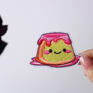 May include: A pink and yellow embroidered iron-on patch of a cartoon pudding with a happy face and pink frosting.