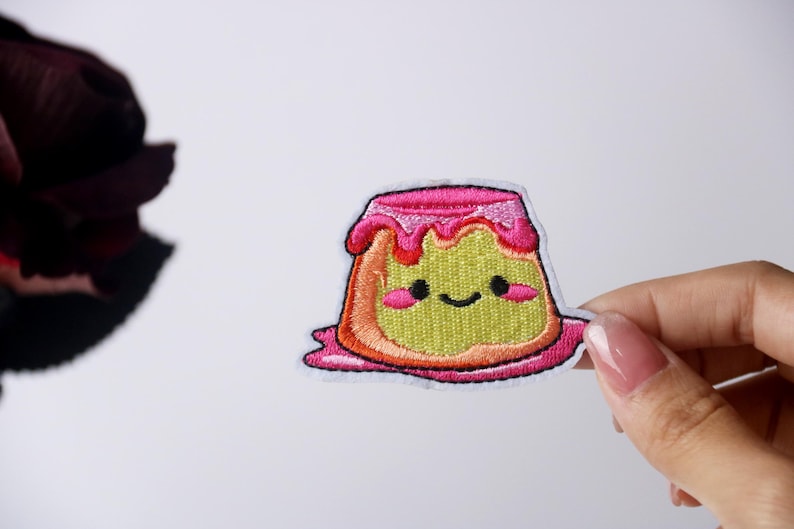 Peut inclure: Un patch brod&eacute; &agrave; repasser en forme de pudding de dessin anim&eacute; avec un visage souriant et un gla&ccedil;age rose.