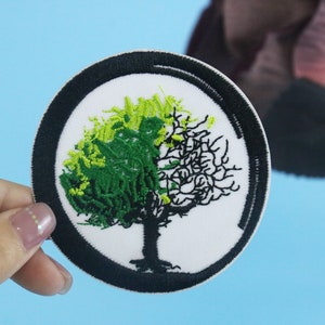 May include: A white embroidered patch with a black circle around the edge. The patch features a stylised tree with green and black leaves. The tree is split in half, with one side green and the other black.