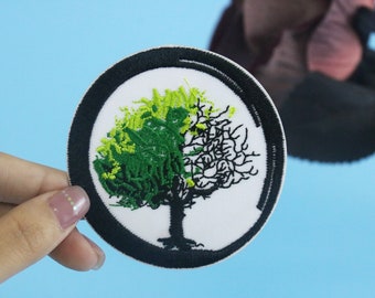 May include: A white embroidered patch with a black circle around the edge. The patch features a stylized tree with green and black leaves. The tree is split in half, with one side green and the other black.