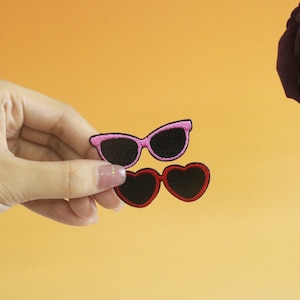 May include: Two embroidered iron-on patches of sunglasses. One pair is pink with black lenses and the other pair is red with black lenses.