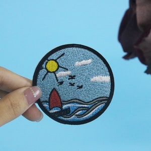 May include: A blue and white embroidered patch with a red sailboat, a yellow sun, and white clouds. The patch is surrounded by a black border.