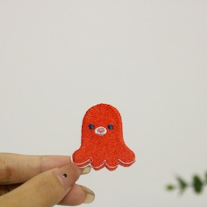 Może przedstawiać: Czerwona haftowana łatka z ośmiornicą z białymi konturami i niebieskimi oczami. Ośmiornica ma różowy nos i przyjazny wyraz twarzy.