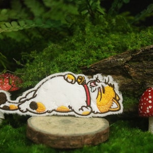 May include: A white, yellow, and brown embroidered patch of a cartoon cat laying on its back with its paws in the air. The cat is wearing a red collar with a bell. The patch is on a small wooden log surrounded by green moss and red and white mushrooms.