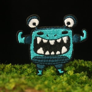 May include: A blue and black embroidered patch of a cartoon monster with a wide open mouth and two raised arms. The monster has large white eyes and a black outline.