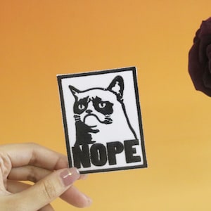 Puede incluir: Un parche termoadhesivo blanco y negro con una imagen de un gato gruñón y la palabra "NOPE" en letras negras.