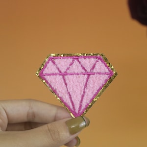 May include: Pink diamond-shaped patch with a gold glitter outline. The patch is made of fuzzy fabric.
