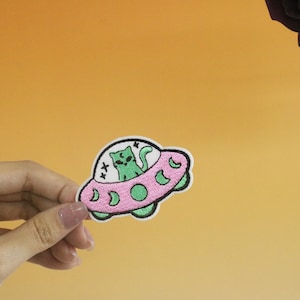 May include: An embroidered patch featuring a cat riding in a UFO. The UFO is pink with a white outline and a green interior. The cat has green eyes and a white star above its head.