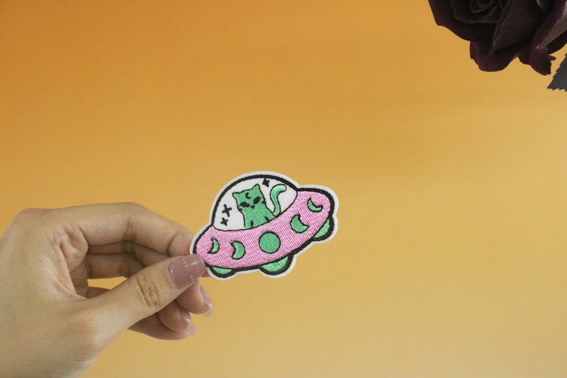 May include: An embroidered patch featuring a cat riding in a UFO. The UFO is pink with a white outline and a green interior. The cat has green eyes and a white star above its head.