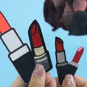 Op de afbeelding: Vier geborduurde strijkbare patches van rode lipsticks in verschillende stijlen. De lipsticks zijn in verschillende tinten rood en hebben zwarte contouren.