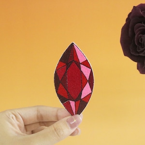 May include: A red and pink gemstone patch with a geometric design. The patch is held in a hand against a light orange background.