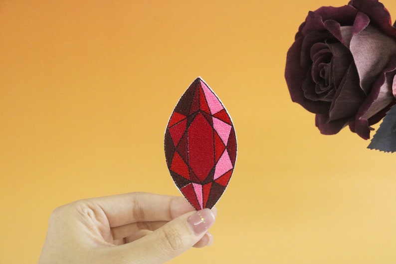 May include: A red and pink gemstone patch with a geometric design. The patch is held in a hand against a light orange background.