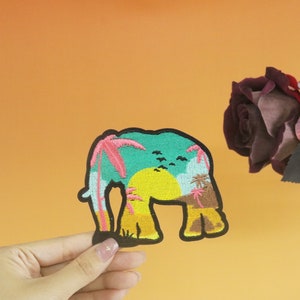 May include: A colourful embroidered patch featuring an elephant silhouette with a tropical sunset scene inside. The patch is green, blue, yellow, pink, and brown.