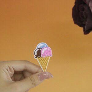 Op de afbeelding: Een geborduurde strijk-op patch met een cartoon-ijsje met drie bollen ijs in roze, bruin en blauw.