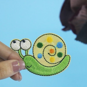 Peut inclure: Un patch brodé en forme d'escargot vert et jaune avec des points colorés sur sa coquille.