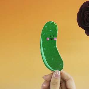 May include: An embroidered patch of a smiling pickle with pink cheeks, in green.