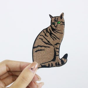 Op de afbeelding: Een geborduurde strijk-op patch van een tabby kat die zit met groene ogen.