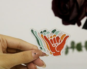 Può includere: Un toppa ricamata colorata da stirare con un designo a strati di una mano che fa il segno shaka, un gesto hawaiano popolare di pace e buone vibrazioni. La toppa è fatta di tessuto bianco con ricami verdi, arancioni e rosa.