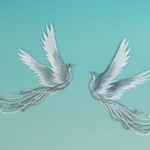 May include: Two silver embroidered phoenixes with long, flowing tails against a light blue background.
