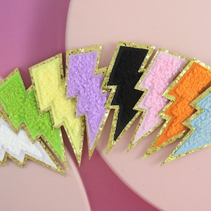 Puede incluir: Un conjunto de diez parches en forma de rayo con ribete de purpurina dorada. Los parches están hechos de tela afelpada en varios colores, incluyendo blanco, verde claro, amarillo, lavanda, negro, rosa, naranja y azul claro.