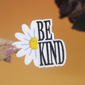 May include: White embroidered daisy with a yellow center and blue petals, partially covering the words "BE KIND" in black embroidery.
