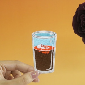 May include: An embroidered iron-on patch of a glass of iced coffee. The glass is clear with a blue liquid and a brown coffee color. The ice is red and white.