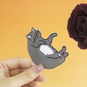May include: A gray and white embroidered cat patch with a white patch on its belly. The patch is being held by a hand against a yellow background.