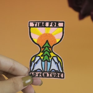 May include: An embroidered iron-on patch with a pink, yellow, and blue hourglass design. The hourglass is filled with a sun, a green pine tree, and blue mountains. The text "TIME FOR ADVENTURE" is written on the patch.