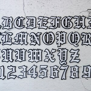 May include: White letters and numbers in a black outline, in a gothic font style. The letters are A through Z and the numbers are 0 through 9.