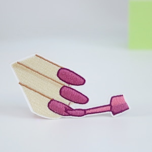 May include: An embroidered patch depicting a hand painting fingernails with pink nail polish. The patch is beige with brown lines and pink details.