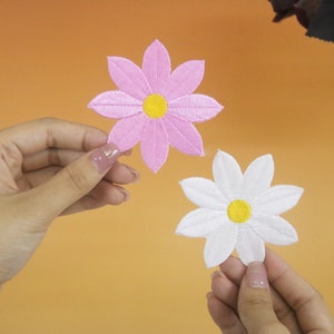 May include: Two embroidered fabric flower patches, one pink and one white, with yellow centres. The patches are held in the hands of two people.