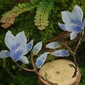 Könnte beinhalten: Zwei blaue gestickte Magnolienblüten-Patches mit braunen Stielen und grünen Blättern. Die Patches befinden sich auf einer Holzoberfläche mit grünem Moos.