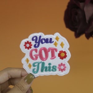 Pode incluir: Etiqueta branca bordada com o texto "You Got This" em letras coloridas. A etiqueta é decorada com pequenas flores e estrelas bordadas.