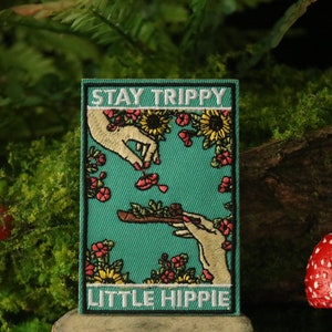 Op de afbeelding: Een geborduurde patch in tealblauw met de tekst "STAY TRIPPY LITTLE HIPPY" in wit. De patch toont twee handen, één houdt een pijp vast en de andere laat bloemen vallen. De achtergrond is gevuld met bloemen en zonnebloemen.