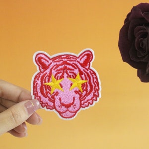 May include: Pink embroidered iron-on patch of a tiger with yellow star-shaped eyes.