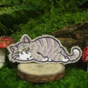 May include: A gray and white embroidered cat patch with a grumpy expression, lying on a small wooden log surrounded by green moss and red and white mushrooms.