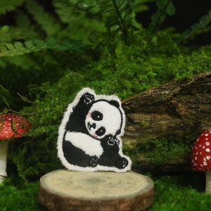 Peut inclure: Un patch brodé en forme de panda noir et blanc avec un fond blanc et une fourrure noire. Le panda est assis avec ses pattes levées et a une expression mignonne sur son visage. Le patch est posé sur une petite bûche de bois avec de la mousse verte autour.